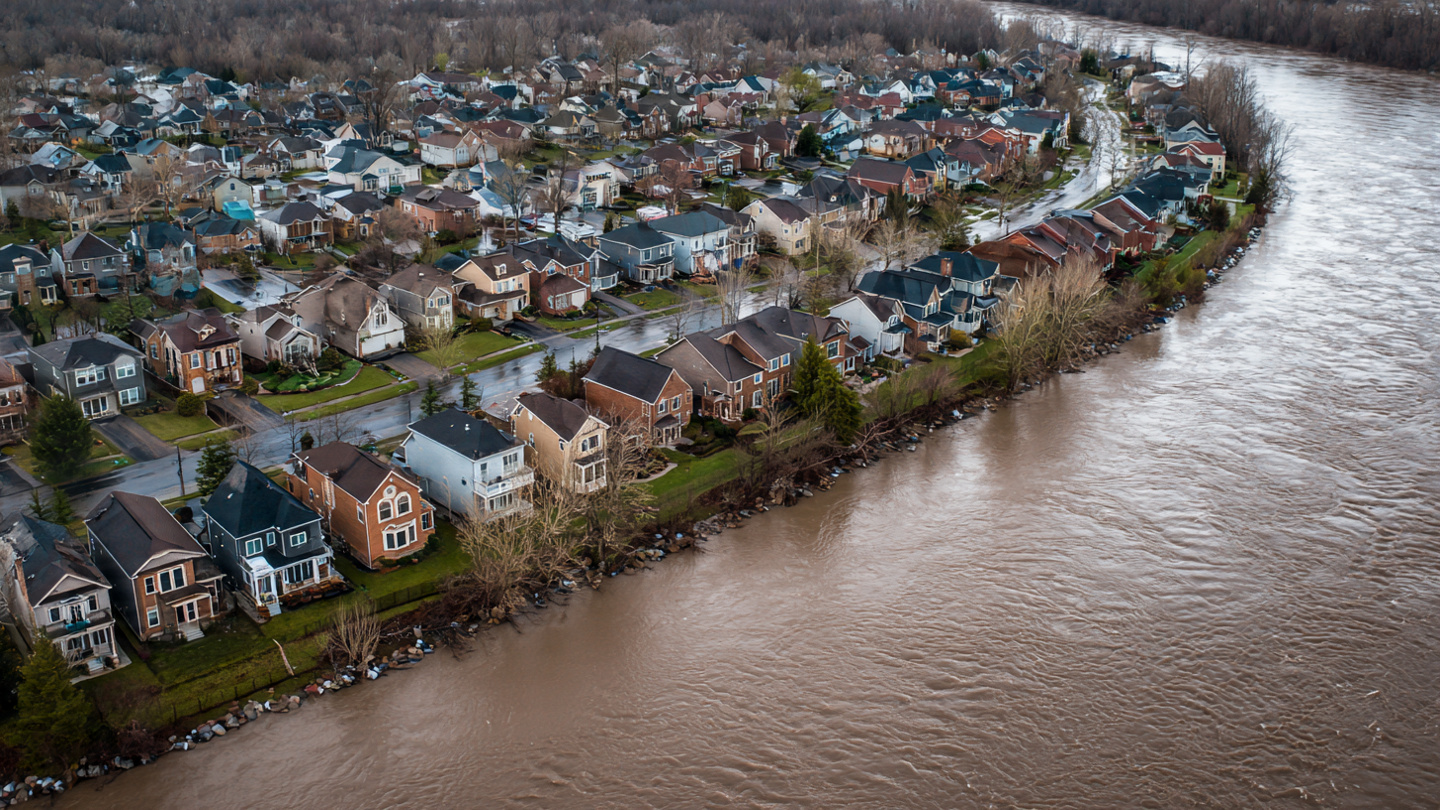 Aerial view of a suburban neighborhood next to a swollen river with sandbags visible